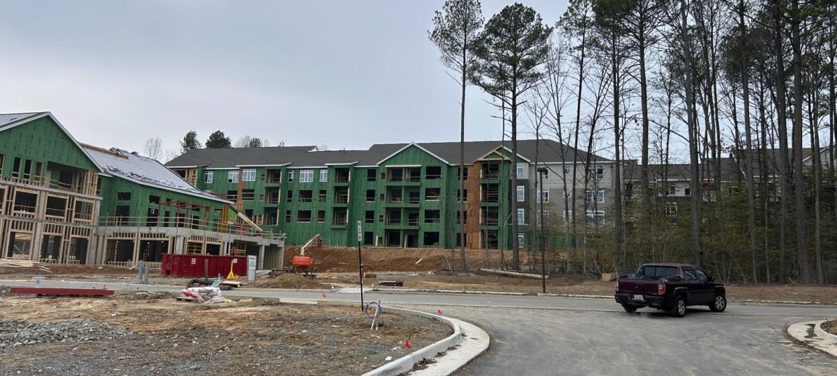 Apartment buildings nearing completion next to wooded area with dirt path and construction equipment.